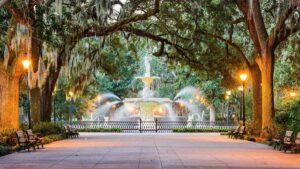 Wide shot of the fountain spraying water at Forsyth Park with a grove of trees leading up to it with black iron light poles lining the sidewalk in Savannah, Georgia, USA